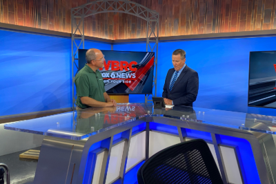 Image of two men in a news studio with a monitor behind them that says 